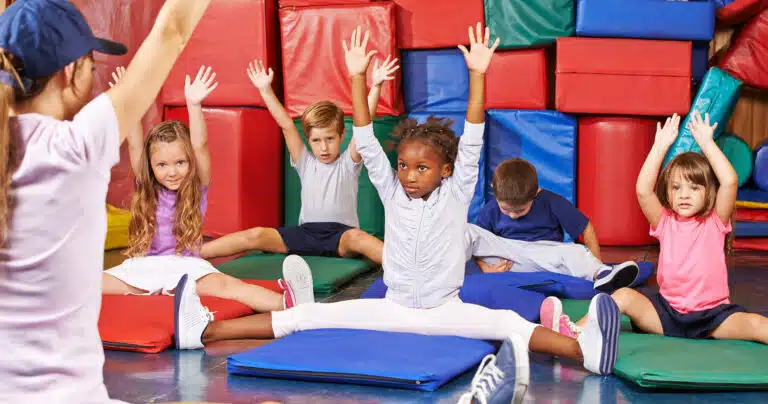 Children facing teacher, with arms raised in gymnastics class